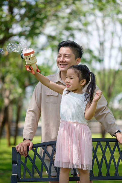 Joyful little girl standing on a bench, blowing bubbles while her dad stands proudly behind her.