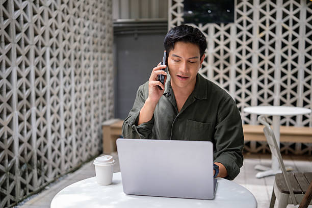 istock Young man seated at a cafe table using a laptop and speaking on a smartphone, representing remote communication and business multitasking. 2238962656