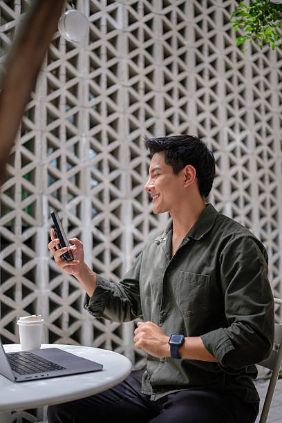 istock Smiling man holding a smartphone and chatting with someone, seated at a table with coffee and a laptop, representing connection and modern communication. 2238962696