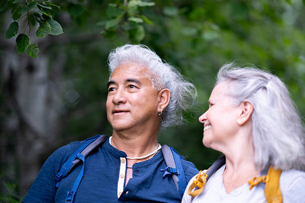 A smiling gray-haired couple with backpacks enjoys a serene moment in a green forest, showcasing companionship, outdoor adventure, and healthy aging during a nature hike.