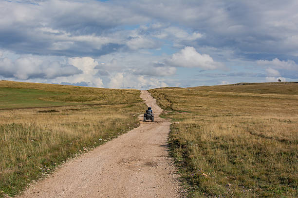 Two people ride ATVs across a mountainous karst plateau on a partly cloudy autumn day