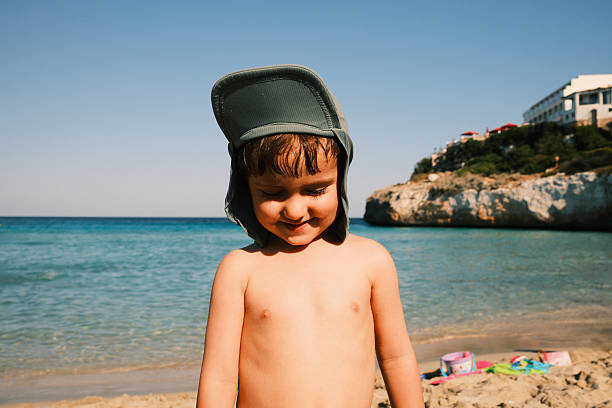 Young boy in a sun hat smiling shyly at the beach, half-length portrait on a sunny Mediterranean day by the sea
