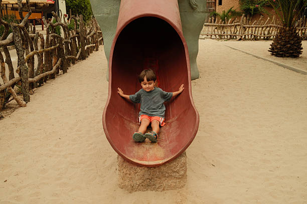 Happy little boy enjoying playtime, sitting at the bottom of an elephant-shaped slide in a sandy playground