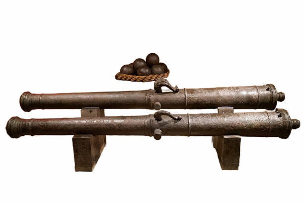 Two antique cannons are prominently displayed in a museum setting, showcasing their detailed craftsmanship. Nearby, a basket containing cannonballs adds to the historical context of the exhibit.