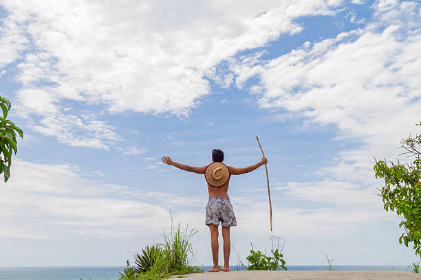 Shirtless young muscle latin surfer male standing on a hilltop with arms wide open, holding a stick, wearing a straw hat on his back, admiring the vast ocean and cloudy sky during summer vacation in Rio de Janeiro, Brazil.He's wearing  a straw hat and light blue board shorts.