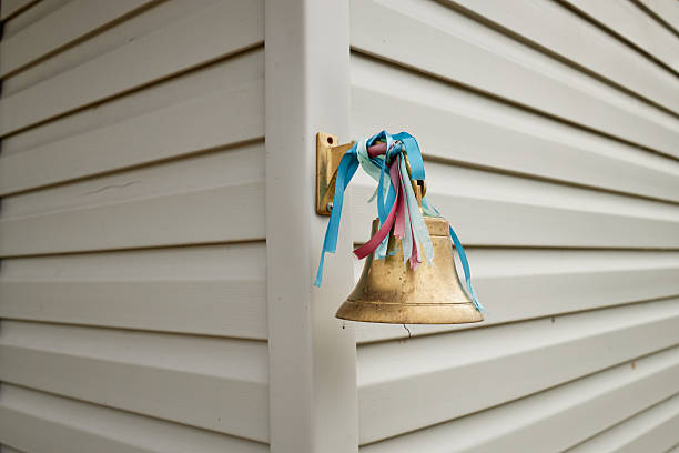 Brass bell with blue and pink ribbons hanging on exterior wall of building, metal fixture mounted on siding, corner of structure visible, no people present in scene