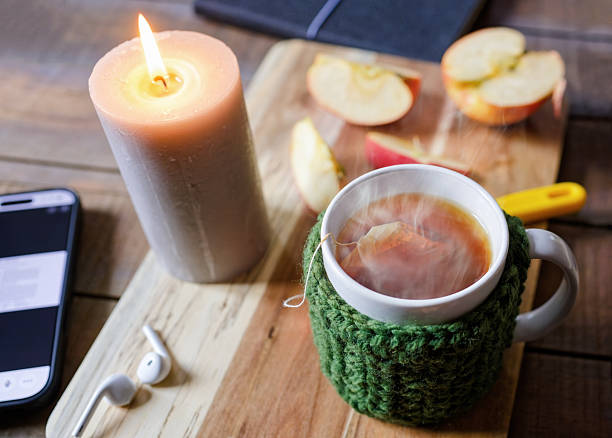 A lit candle, steaming mug of tea with a knitted cozy, sliced apple, a phone and a diary sit on a wooden surface, creating a cozy and relaxing evening scene. High angle view - Concepts and ideas, healthy lifestyle