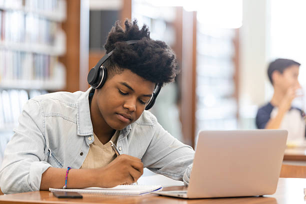 istock Young student with headphones using a laptop and writing notes in a library. 2239004573