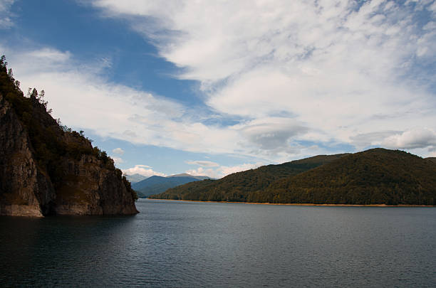 Wide view of a tranquil mountain lake or reservoir surrounded by dramatic natural features. A massive, shadowed rock face rises sharply on the left, contrasting with the lush, forested slopes stretching into the distance under a dramatic, cloudy sky.