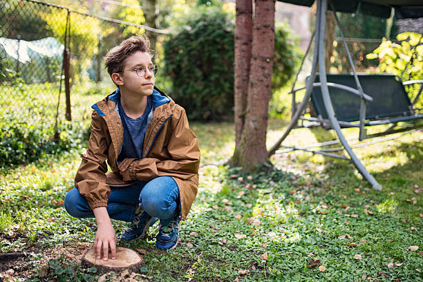 Teen with short hair and glasses crouching on a grassy lawn, one hand gently placed on a tree stump. Dressed in a brown jacket with blue lining, a blue shirt, jeans, and sneakers, they appear quietly absorbed in the moment. \n\nShot with Canon R5