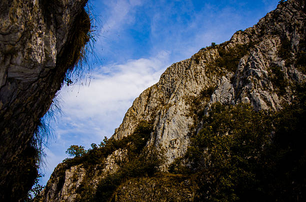A low-angle, dramatic view looking up through a mountain gorge. Massive, steep limestone cliffs dominate the frame, covered sparsely with scrub vegetation. The bright blue sky and clouds provide a striking contrast to the rough, textured rock walls.