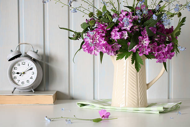 Bouquet of purple flowers in a jug next to an old alarm clock and book on a table
