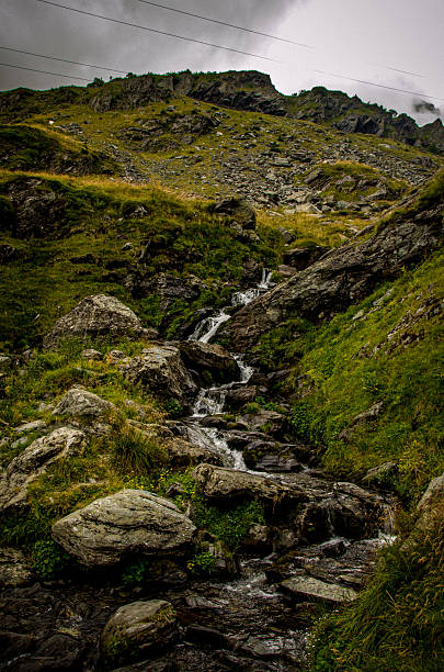A dramatic vertical view of a mountain stream rapidly flowing down a steep, boulder-strewn slope. Lush green and yellow grass covers the rugged hillsides. The atmosphere is moody and cool due to the dense gray, overcast sky. Visible power lines cross the upper frame.
