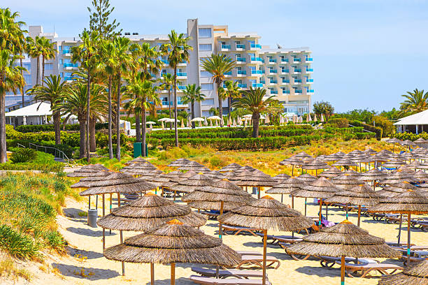 Nissi Beach resort in Ayia Napa, Cyprus, amidst lush greenery and a sandy beach lined with straw umbrellas. Tranquil sunny day at this popular Mediterranean tourist destination, perfect for relaxation