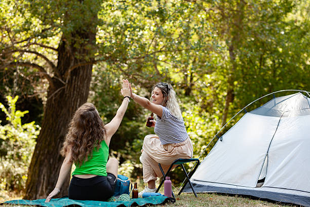 istock Two women are sharing a joyful interaction while camping in a lush forest. One sits on a chair, and the other is seated on a blanket, both engaging in conversation and laughter 2239077471