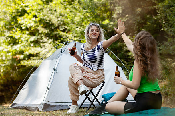 istock Two women relax at a campsite, laughing and sharing drinks while enjoying nature. Their tent is set up nearby, showcasing a typical summer camping experience 2239077496