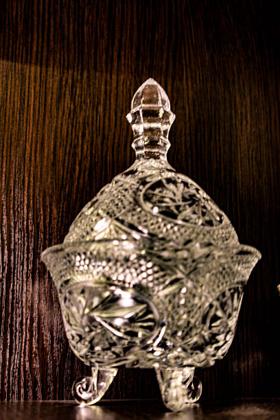 A single crystal candy dish or sugar bowl with exquisite carving, a spire lid, and three elegant legs stands on a dark wooden shelf.