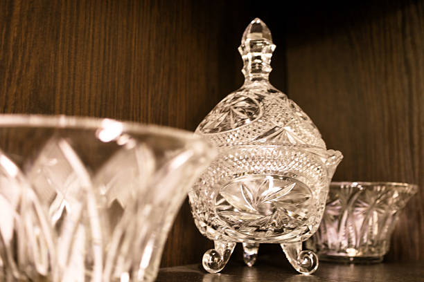 istock Crystal glassware items with decorative carving stand on a dark shelf, among which a candy dish or sugar bowl on legs with a spire lid stands out. The close-up shot shows the light reflections and luxury of vintage glass against a blurred wooden backgroun 2239093217