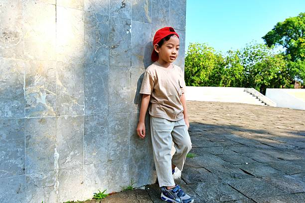 A young boy leans against a white wall outdoors, dressed in casual beige outfit and red cap, under warm natural sunlight.