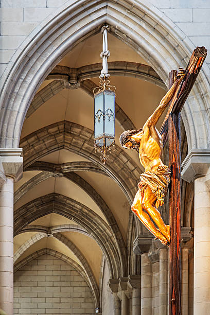 Catedral de Santa Maria la Real de la Almudena, Madrid, Spain, Europe, September 12, 2025: Dramatic close-up of a crucifix sculpture depicting Jesus Christ on a wooden cross, prominently displayed inside a Gothic-style cathedral in Madrid, Spain. The figure is illuminated by ambient light, emphasizing the emotional intensity and craftsmanship of the religious artwork. Architectural details such as pointed arches, ribbed vaults, and a hanging lantern contribute to the solemn and reverent atmosphere.
