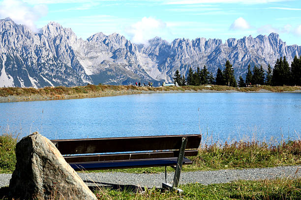A quiet spot features a wooden bench by a peaceful pond, framed by majestic mountains in the background. The sunlight reflects off the water, adding to the beauty.