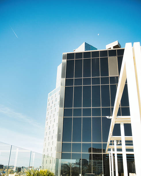 A stylish office terrace featuring a cozy patio with puffs, large white pots with palm trees, and a wooden floor. The view includes a modern building with black transparent windows, a clear blue sky, and the moon visible in the background on a sunny day. Perfect for themes of modern architecture, outdoor relaxation, and urban design.