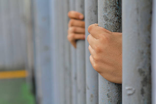 istock prisoner's hands behind bars with black background 2239139175