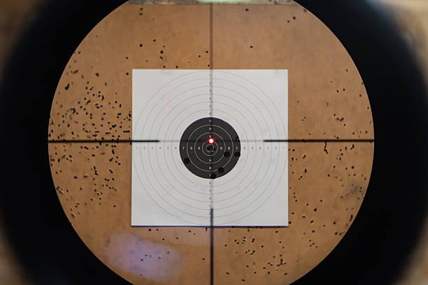 A first-person view through a rifle scope with a red dot sight aimed at a paper bullseye target outdoors.Close up photo