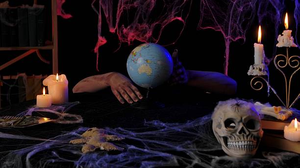 Scary zombie hands holding a globe. A dark room with burning candles, a skull on the table, and creepy cobwebs. Suitable for Halloween themes, spooky decorations, and holiday media publications.