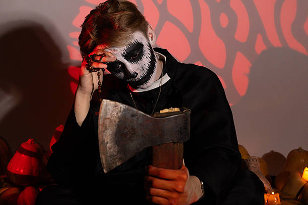 Man with face painted in striking black and white makeup, staring directly at the camera with intense eyes against a dark background