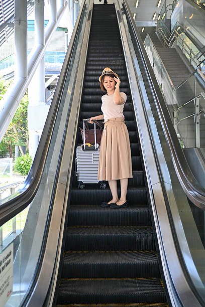 An Asian woman with luggage holds her head in pain as she rides the escalator, showing signs of travel exhaustion.