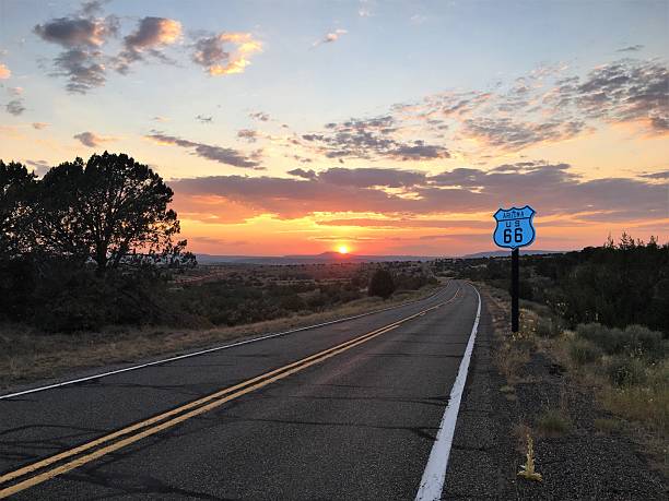 A scenic stretch of Historic U.S. Route 66 captured during golden hour. The iconic blue-and-white road sign stands prominently beside the empty highway, which disappears into the horizon beneath a sky painted with warm sunset hues and scattered clouds. Natural vegetation and trees line the road, evoking a sense of nostalgia, freedom, and timeless Americana. Ideal for themes of travel, history, solitude, and the open road.