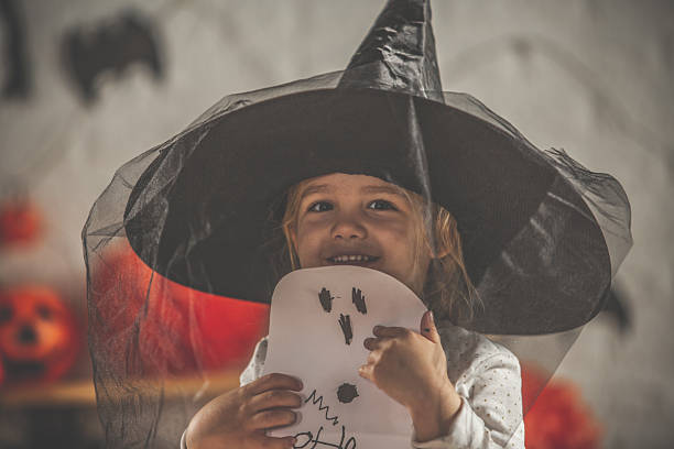 A young girl wearing a tall black witch hat holds a paper drawing and smiles warmly, creating a charming Halloween moment with soft lighting and festive background hints.