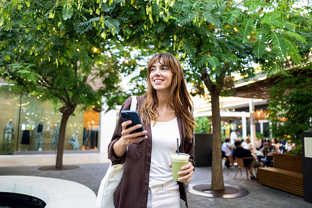 istock Smiling woman enjoying an iced matcha latte, browsing smartphone while walking through an urban shopping area with trees 2239183271
