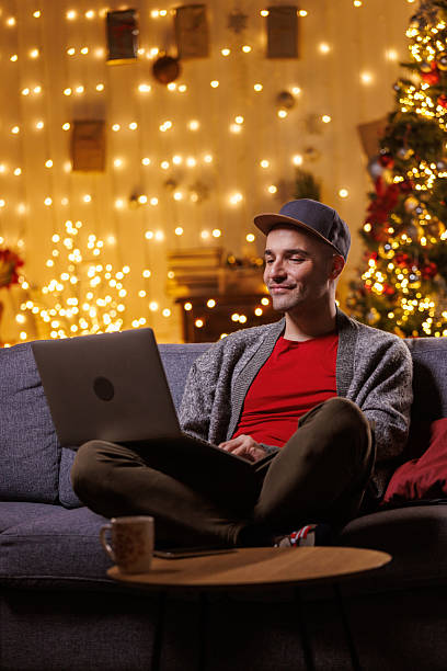istock Man relaxing on couch with laptop amid cozy Christmas lights and tree decorations 2239186846