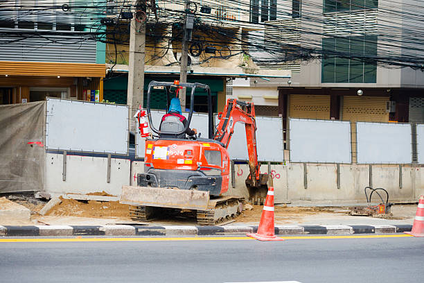 Thai worker in small backhoe machine is working on Rama IX Road in Bangkok. Man is working for repairing works below overpass road.  View over street from opposite side. Man is working in center of street. In background are buildings