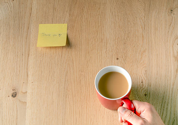 A red coffee mug of coffee on the wooden table and a yellow sticky note on the side that says Thank you. A person is holding the mug