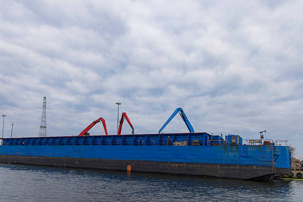 A large blue cargo barge with multiple crane arms sits on still water, set against a cloudy sky. Industrial and maritime elements convey transport, loading, and shipyard activity.