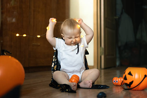 istock toddler sits on the floor, wearing a Halloween cape, joyfully playing with string lights and surrounded by pumpkin decorations. The child displays a playful and joyful expression. 2239220716