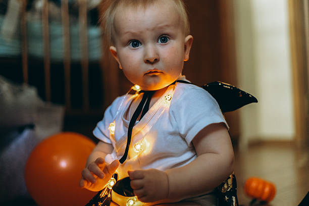 istock Baby dressed in a Halloween costume sits with glowing string lights around their neck, surrounded by a festive atmosphere. The child's expression is curious and innocent, capturing the essence of a playful Halloween setting. 2239220733