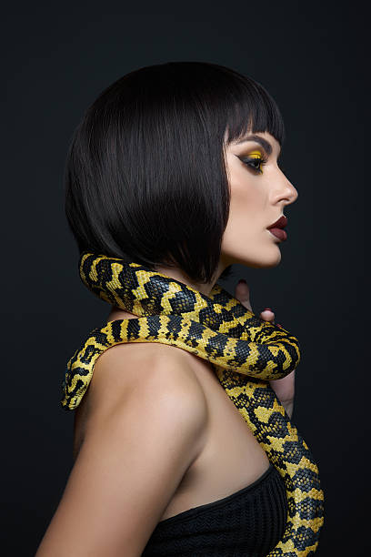 istock In a dramatic dark setting, a woman with a sleek black bob hairstyle poses confidently. She has bold makeup, and a yellow and black striped snake rests around her neck, creating a striking contrast 2239224654