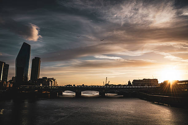 Sunset over the River Thames in London, England, with the dramatic sky casting warm golden light on the water. The silhouette of modern buildings and bridges, including Blackfriars Bridge, forms a striking contrast against the glowing horizon.