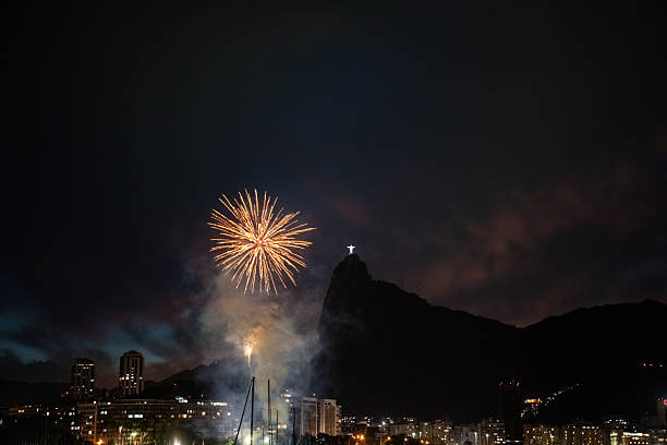 Nighttime photograph taken in Urca, Rio de Janeiro, showing fireworks exploding near Corcovado Mountain. The scene captures the festive glow of the fireworks against a dark sky with faint purple and blue tones.