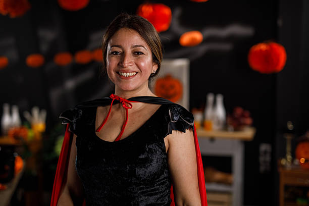 A woman in a witch costume with a black dress and hat poses against a textured background, celebrating Halloween.