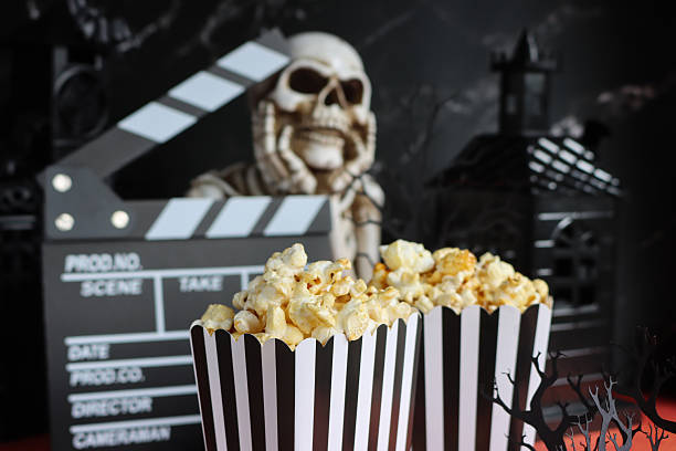 Close-up image black and white striped popcorn cartons, film snack food tubs, open film clapperboard, 3D paper craft tree, model skeleton chin in hands, background of spooky houses, focus on foreground, Halloween scary film concept stock photo