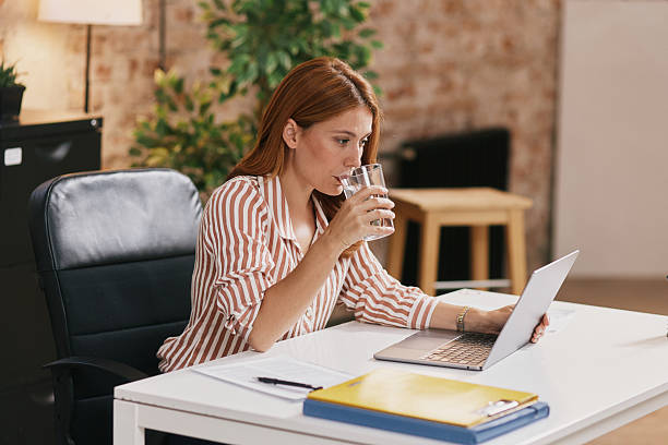 istock Adult Woman Working at White Desk, Drinking Water While Using a Laptop in a Modern Office 2239264200