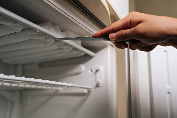 istock Hand holding knife scraping ice from inside of freezer compartment, showcasing need for defrosting to maintain efficient cooling. 2239265747