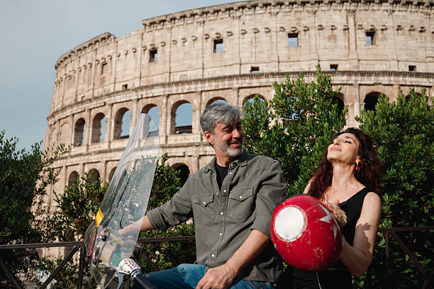 A mature couple is getting ready for a motorcycle ride in Rome, Italy. The man sits on a scooter while the woman holds a red helmet, preparing for a scenic adventure near the iconic Colosseum. A perfect moment showcasing senior travel, freedom, and tourism in Italy.