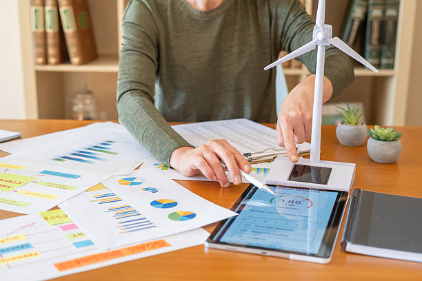 istock Woman managing renewable energy project, analyzing tablet data, pointing at wind turbine model while reviewing charts and graphs on desk, planning sustainable business and clean tech 2239289394