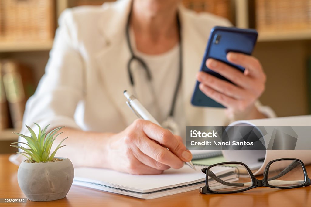 Médico con un estetoscopio que trabaja desde una oficina en casa, tomando notas en papel mientras sostiene un teléfono inteligente, representando la telemedicina moderna y la práctica general - Foto de stock de Adulto libre de derechos Médico con un estetoscopio que trabaja desde una oficina en casa, tomando notas en papel mientras sostiene un teléfono inteligente, representando la telemedicina moderna y la práctica general - Foto de stock de Adulto libre de derechos